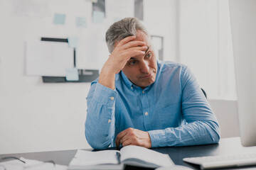 Fototapeta premium handsome tired gray-haired man sits in his office holding hand near head looking upset while working on business project , stress at workplace concept