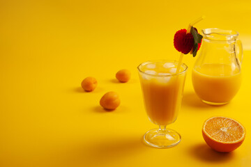 Glass with cold orange juice with ice, straw, apricots, orange half and glass jar. Yellow background. Copy space.