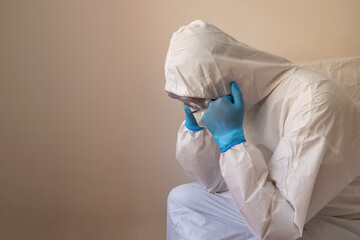 Fototapeta premium Doctor with anti-virus protective suit sitting on the bed with his hands covered in disposable gloves under his chin