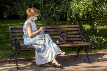 Woman with a medical mask on her face is sitting on a park bench knitting wool clothes on knitting needles and watching lessons or teaching knitting through a laptop. Distance learning