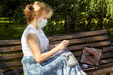 woman with a medical mask on her face is sitting on a park bench knitting wool clothes on knitting needles and watching lessons or teaching knitting through a laptop. Distance learning