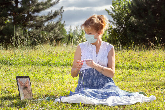 woman with a medical mask on her face sits on the grass in a park knitting wool clothes on knitting needles and watches lessons or teaches knitting through a laptop. Distance learning - Powered by Adobe