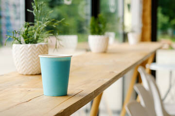 A blue paper cup with hot fresh coffee is standing on a wooden table in a cafe. The interior of the cafe with a Ripsalis cactus (twig) and other green plants. The view from the coffee shop.