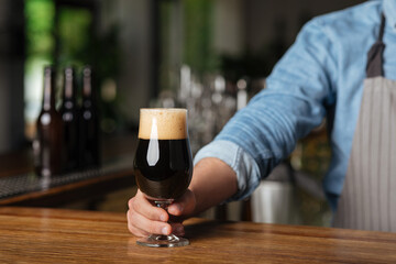 Serving client with drinks. Barman in apron gives glass of dark beer with foam on wooden bar