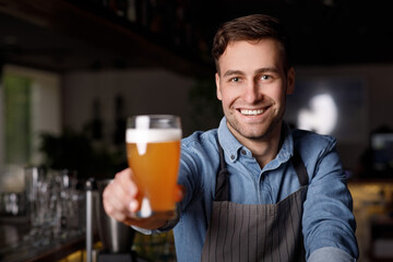 Work in pub. Smiling handsome barman in apron holds out glass of beer