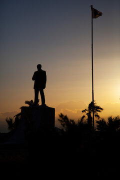 Image shows the silhouette of a Mustafa Kemal Ataturk monument, and a tall flag post where Turkish flag is flying at full mast at sunrise. There are spectacular views of clouds, sky as well as trees 