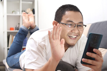 Asian man doing a video call at home, smiling to camera and waving
