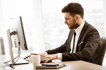 Business concept. Successful young businessman at work. Manager sitting at the office table and working on computer. Busy man in suit indoors on glass window background