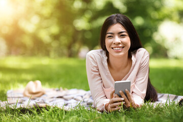 Dreamy Asian Girl Relaxing On Plaid In Park With Smartphone In Hands