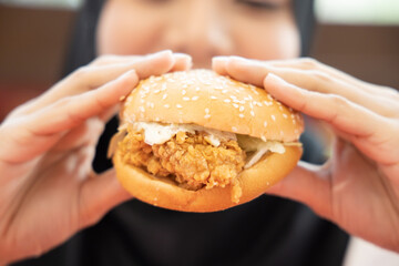 hungry islamic woman looking, eating halal fried chicken burger; concept of delicious halal food, fast food, health care, eating habit, crispy tasty fried chicken burger; asian muslim woman model