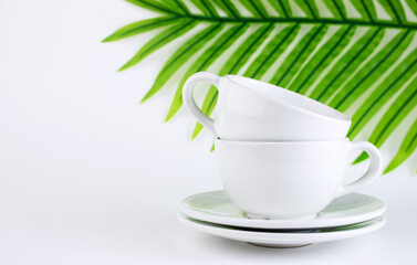White mug and saucer on a white background, near palm leaves. A set of porcelain or ceramic dishes. Copy space