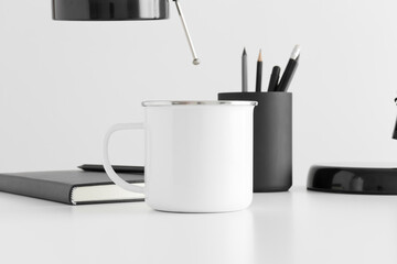 Enamel mug mockup with a lamp, notebook and workspace accessories on a white table.