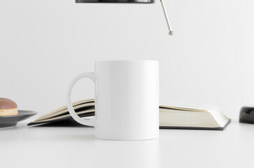 White mug mockup with a donut, book and a lamp on a white table.