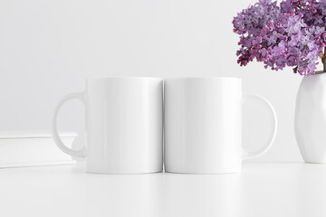 Two white mugs mockup with a book and a bouquet of lilac in a vase on a white table.