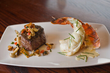 Surf ‘n Turf, lobster tail and steak entree. Filet mignon topped with chanterelle mushrooms & Maine lobster tail topped w/ drawn butter served on white dinner plate. Classic steakhouse menu favorite.