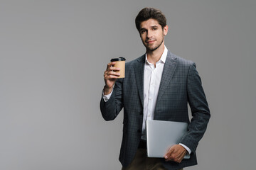 Photo of pleased unshaven businessman drinking coffee takeaway and holding laptop