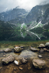 Beautiful view of foggy mountains cover by dark clouds and green forest with a reflection in a lake. Stony shore. Morskie Oko. Marine Eye. High Tatras, Zakopane © WellStock
