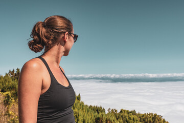 Young brunette woman with sunglasses sitting at the edge of the mountain over the clouds