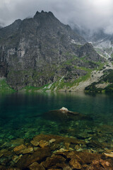 Fototapeta na wymiar Scenic view of foggy mountains cover by dark clouds and green forest with a reflection in a lake. Stony shore. Morskie Oko. Marine Eye. High Tatras, Zakopane
