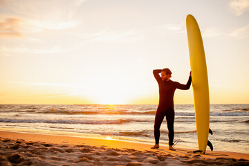 Young bearded surfer in wetsuit with yellow surfing longboard on a ocean coast at sunset. Water sport adventure camp and extreme swim on summer vacation.
