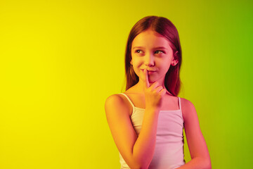 Thoughtful. Little caucasian girl's portrait isolated on gradient studio background in neon light. Concept of human emotions, facial expression, modern gadgets and technologies, sales, ad. Copyspace.