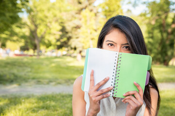 Fototapeta premium A young Indian girl is sitting in the park on the grass, covering her face with a notebook or book. Notebook without inscriptions, with a blank space for any text or advertisement. The time for planni