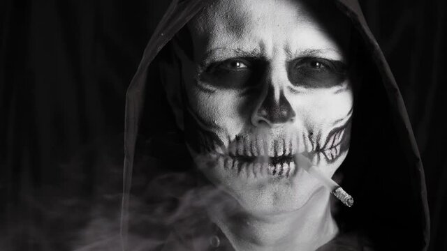 Man with make-up skeleton and black hood on a dark background. Skeleton smokes a cigarette. Smoking kills. Halloween or horror theme. High quality 4k video