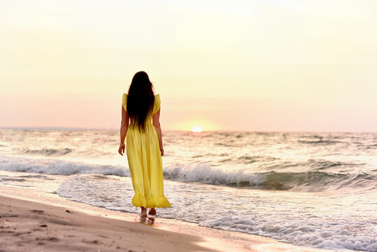 A girl in a long dress with long black hair walks at dawn along a deserted beach along the line of the surf.
