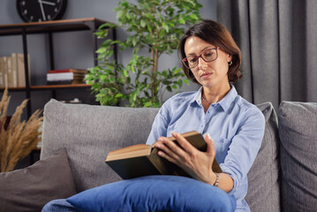 Concentrated lady with brown hair relaxing on cozy sofa with interesting book. Beautiful woman in eyeglasses and casual clothing reading new literature at home.