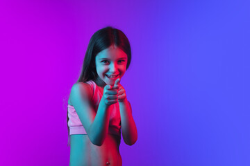 Smiling, pointing on, choosing. Little girl's portrait on gradient pink-blue studio background in neon. Concept of human emotions, facial expression, modern gadgets and tech, sales, ad. Copyspace.