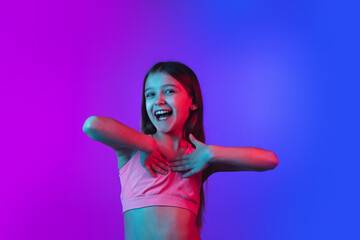 Shy, laughting sincere. Little girl's portrait isolated on gradient pink-blue studio background in neon. Concept of human emotions, facial expression, modern gadgets and tech, sales, ad. Copyspace.
