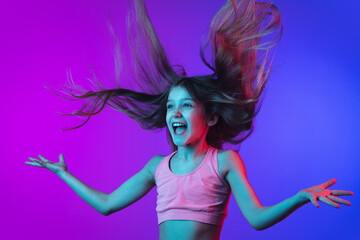 Crazy happy, hair blowing. Little girl's portrait isolated on gradient pink-blue studio background in neon. Concept of human emotions, facial expression, modern gadgets and tech, sales, ad. Copyspace.