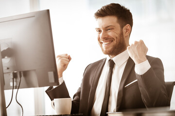 Business succsess concept. Happy smiling young businessman in office gets good news and laughs while work on computer. Man in suit indoors on glass window background - 362914058