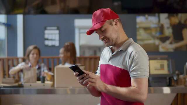 Delivery guy waiting for waiters to pack order and using smartphone in cafe