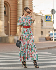 Fototapeta premium Elegant woman in colorful green sundress walking city street. Pretty young girl with wavy dark hair. Fashionable lady