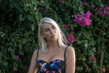 Portrait of young blonde woman in colourful top and green bush with pink flowers behind