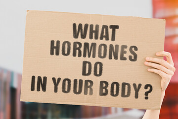 The phrase " What hormones do in your body " on a banner in men's hand with blurred background. Body's cells. Hormone. Biology. Function. Organism. Reproduction. Chemical substances. Hospital. Medical
