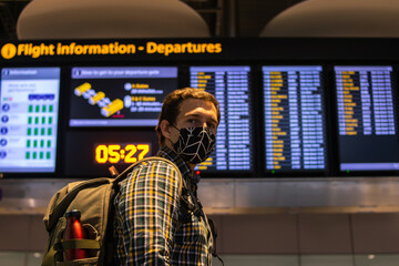 Photo of a guy with a backpack looking at the Departure screen at the airport ready to catch a flight and wearing a reusable face mask.