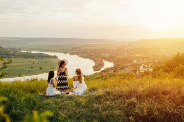 Three female friends in elegant dresses having picnic on the hill  with river on the background. Cozy summer atmosphere. Place for text. Happy young girls friends having fun at picnic. 