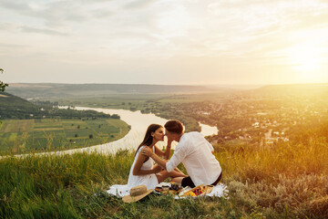 Naklejka na ściany i meble Tender couple sitting on a hill and holding glasses of white wine. Couple on a date sitting on a grass at picnic. Couple in love spending romantic evening together. Copy space.