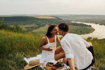 Happy young couple on the hill relaxing at the picnic. Guy and girl on a picnic drink white wine....