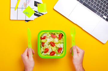 Close up of man hands with food in plastic container.Man eating fresh salad during work.Healthy food concept.Top view, flat lay,yellow background.Healthy snack at office workplace.