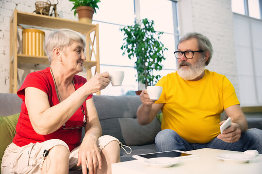 Couple of seniors smiling spending time together being quarantined - caucasians mature and retired man and woman using modern gadgets, talking, drinking tea. Eyes tired of gadget's life. Lifestyle.