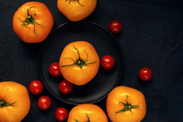Red wet cherry tomatoes and yellow tomato fruits in black plate on a dark textured background. Top view. Space for text.