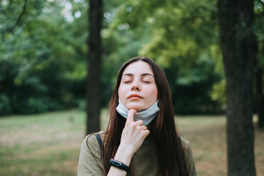Young caucasian pretty woman removes a medical protective mask from her face on nature, surrounded by trees, breathing clean fresh air