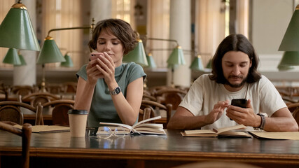 Two young students intently using smartphones while preparing for exams in library. Casual students studying together in university