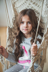 A beautiful young dark-haired girl in gray pajamas relaxes in a swing. Cute brunette girl swinging in a woven chair at home. Looks at the camera. Close-up, soft focus. Vertical photo