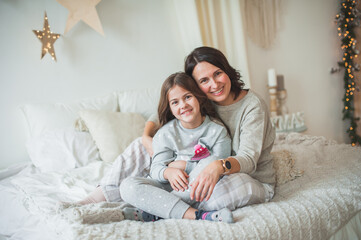 Obraz na płótnie Canvas Beautiful woman and little cute girl, mom and daughter in gray pajamas are sitting on the bed, looking at the camera and laughing. Cozy tender photo, concept, scandy style, soft focus.