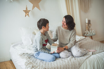 The son makes a surprise to mom and gives her a beautiful box with a Christmas present. Mom smiles. They are sitting on the bed in gray pajamas. Scandi style, light colors, soft focus.