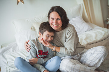 Mom and son are sitting on the bed in the bright bedroom and are smiling happily. Woman hugs teenager boy tightly. The concept of a Christmas morning, scandy style, home comfort, mom and son.